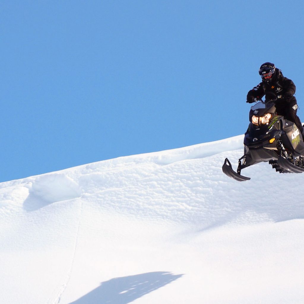 Person riding a snowmobile downhill with clear blue sky. Rovaniemi / Arctic Expeditions - Hibini Mountains Freeride Camp, Wild Nordic Finland @wildnordicfinland
