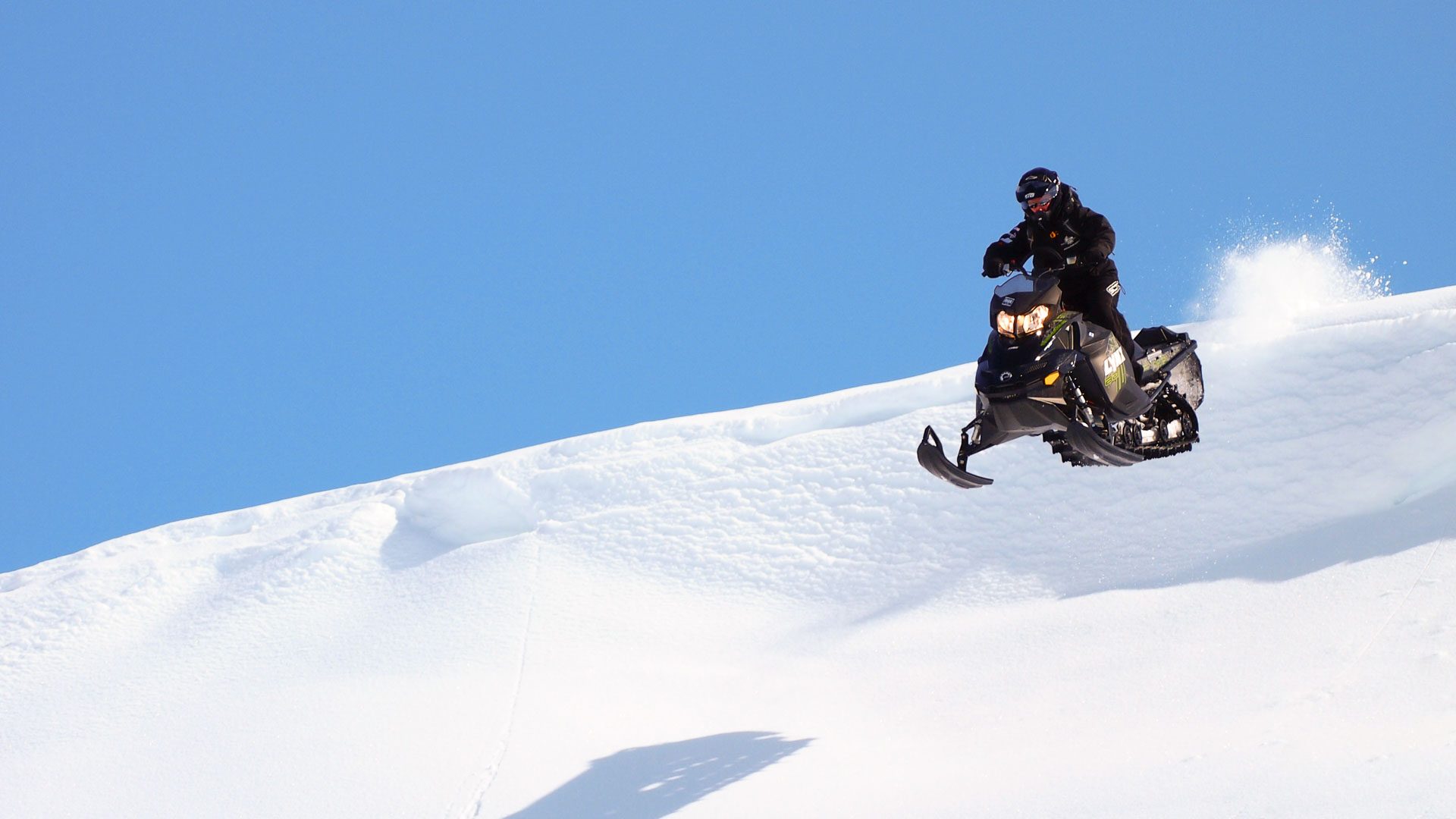 Person riding a snowmobile downhill with clear blue sky. Rovaniemi / Arctic Expeditions - Hibini Mountains Freeride Camp, Wild Nordic Finland @wildnordicfinland