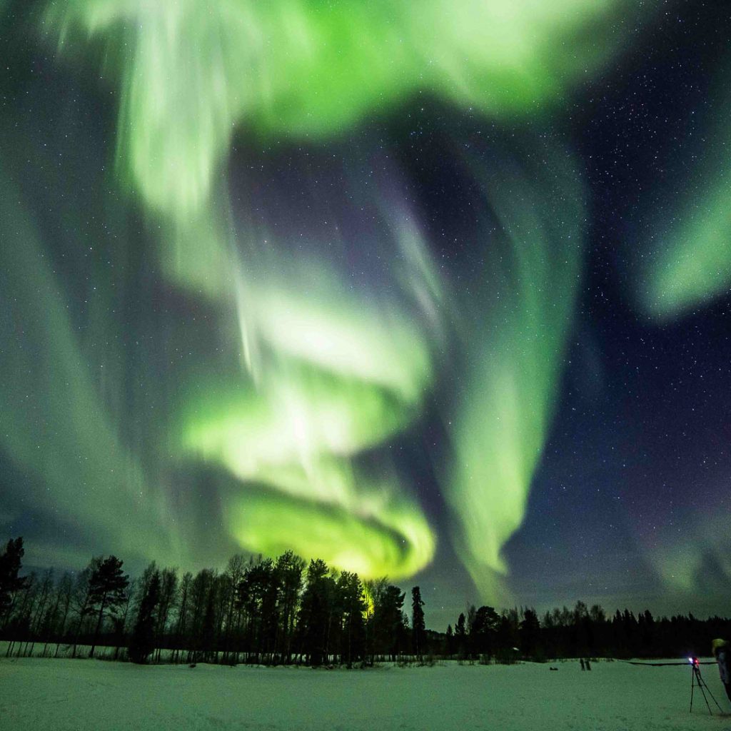 Bright green northern lights and a wooden hut with the fire place burning. Northern Lights Photography Tour, Rovaniemi, Arctic Circle Wilderness Resort, Wild Nordic Finland @wildnordicfinland