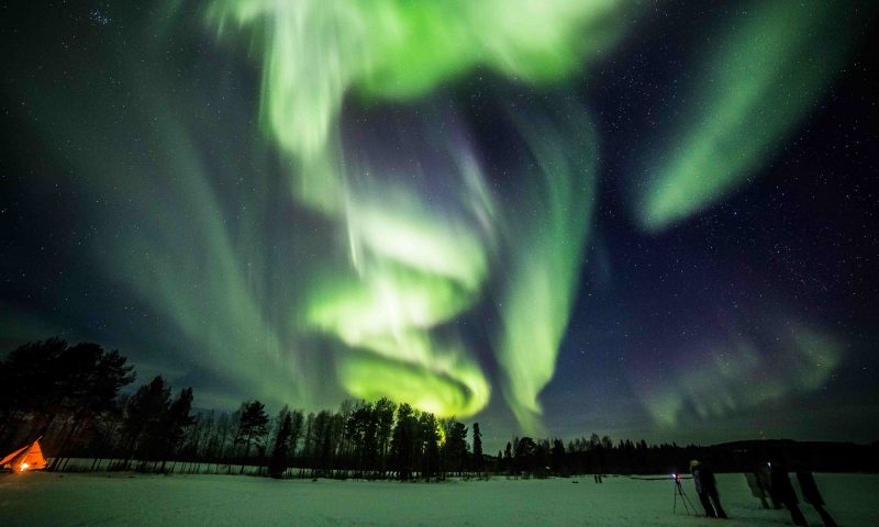 Bright green northern lights and a wooden hut with the fire place burning. Northern Lights Photography Tour, Rovaniemi, Arctic Circle Wilderness Resort, Wild Nordic Finland @wildnordicfinland